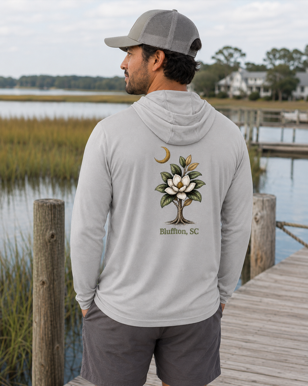 Man wearing a gray hoodie with a tree design and text on the back, standing on a dock by a body of water.