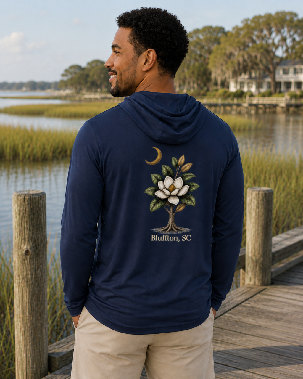 Man wearing a navy hoodie with a tree design and 'Bluffton, SC' text, standing on a wooden dock by a body of water.