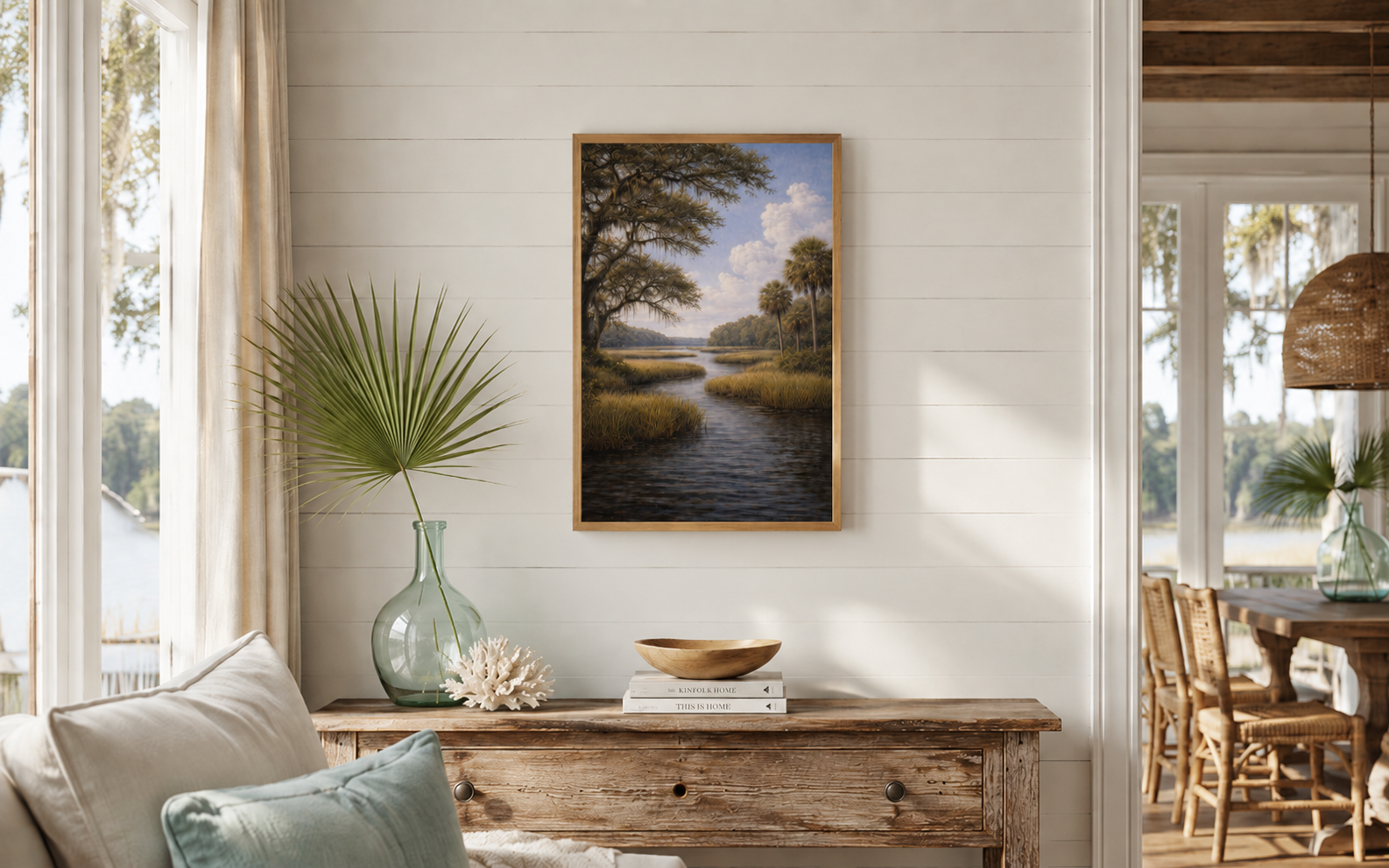 A framed landscape art print depicting a calm river winding through marshlands under a blue sky, styled on a white shiplap wall above a rustic wooden console table in a bright, modern room.