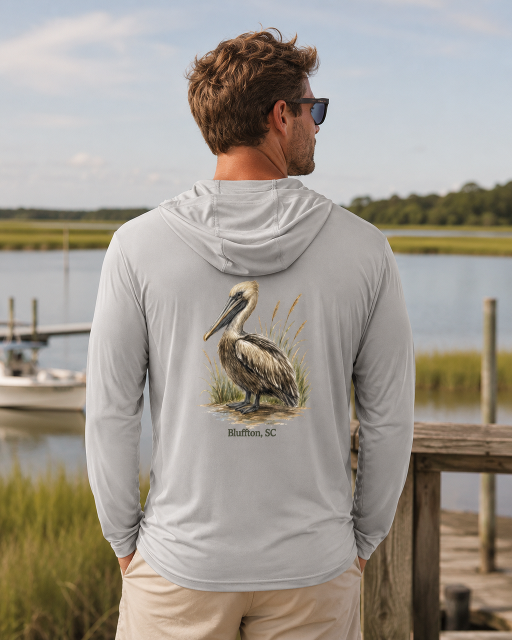Man wearing a gray hoodie with a pelican design, standing by a waterfront.