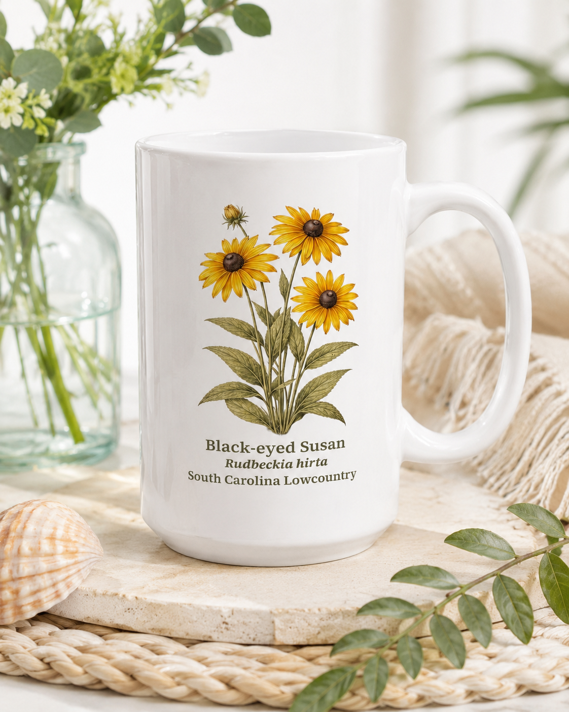 White mug with black-eyed Susan plant illustration and text on a natural setting with plants and shells.