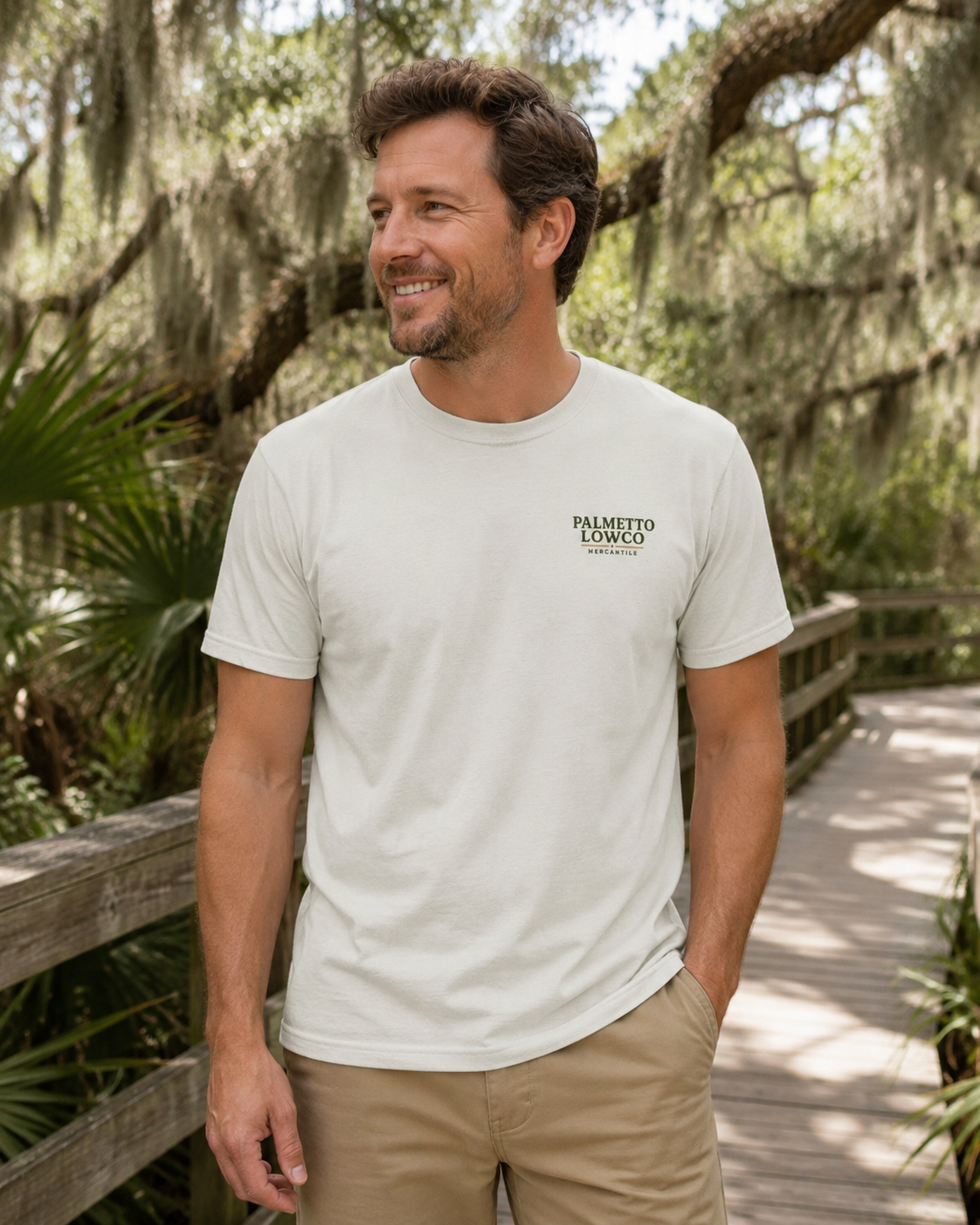 Man wearing a white t-shirt with a logo, standing on a wooden path with greenery in the background