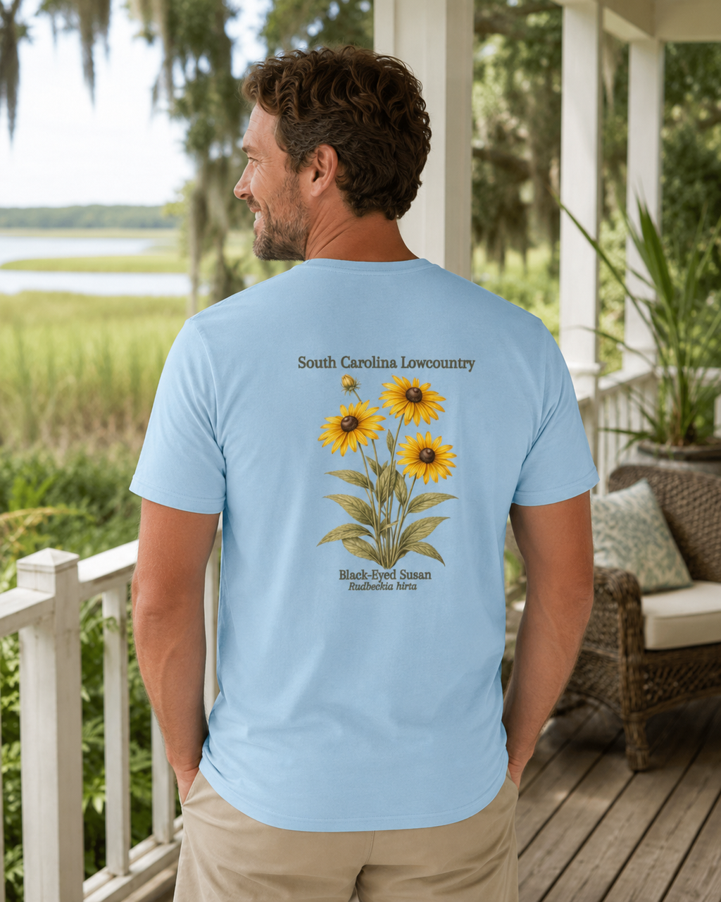Man wearing a light blue t-shirt with back eyed susan design and text, standing on a porch with a scenic background.