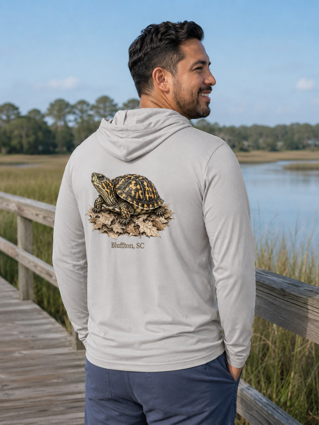 Man wearing a hoodie with a turtle graphic and text, standing by a lake.